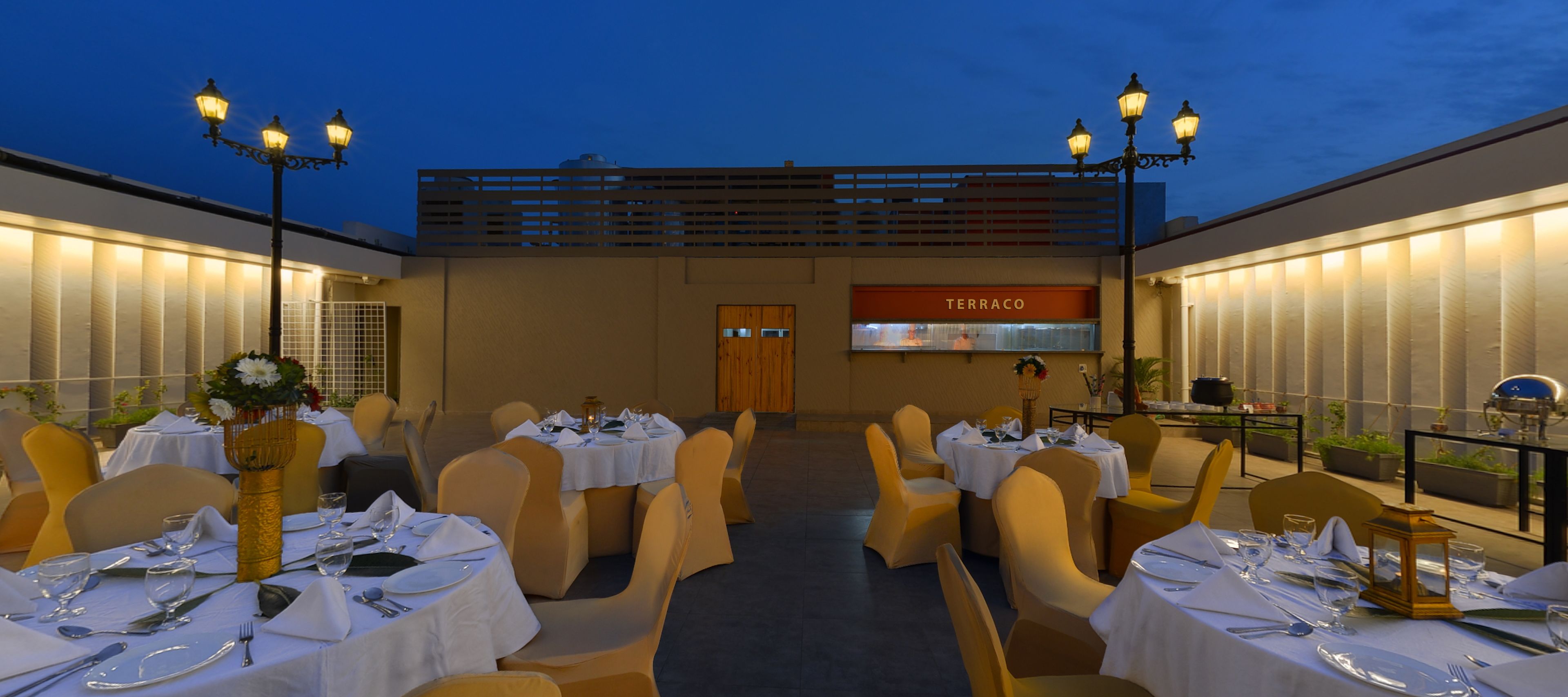 Un área de comedor al aire libre con mesas dispuestas para una comida, un cartel que dice "TERRACO" y una vista del horizonte de la ciudad al anochecer.
