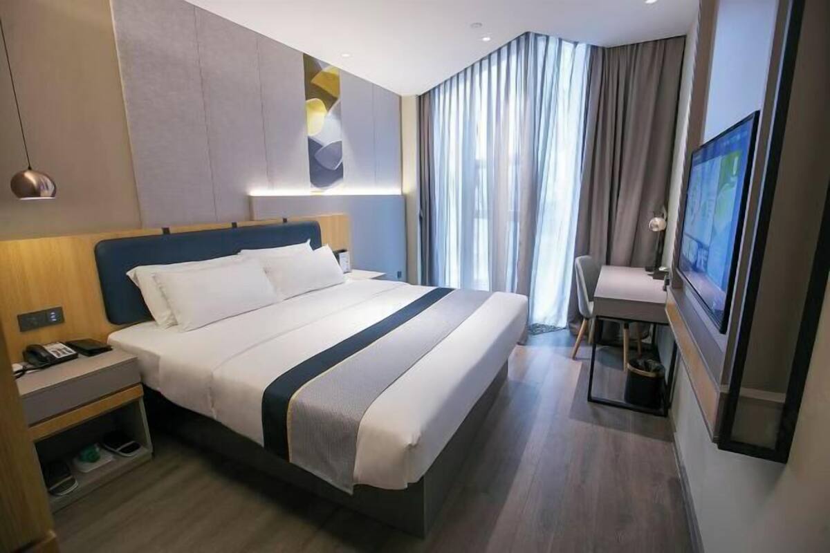 A hotel room with a bed, bedside table, lamp, TV, and a desk with a chair.