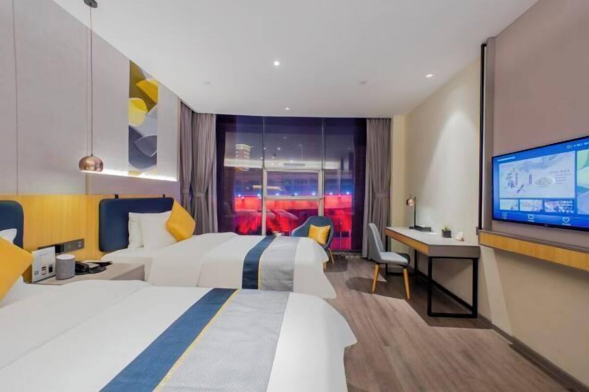 A hotel room with two beds, a desk, a chair, a TV, and a large window with a view of a cityscape.