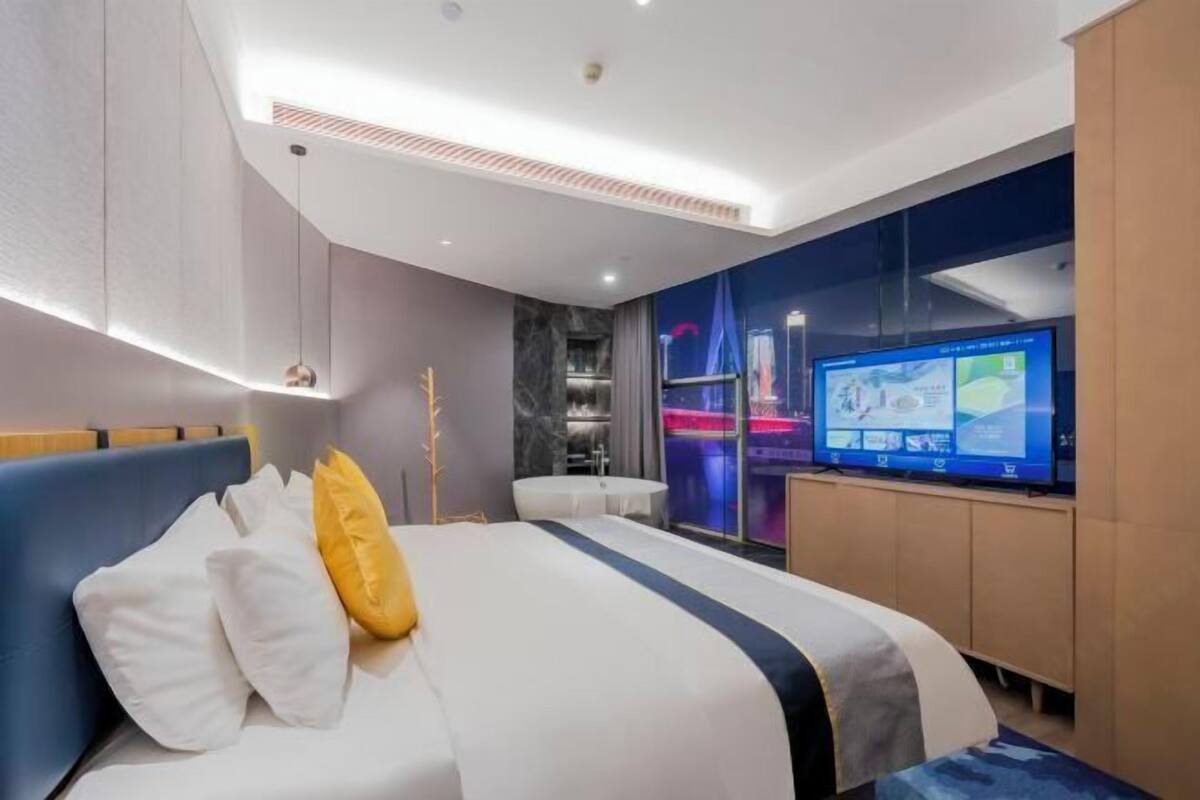 A modern hotel room with a bed, a television, a desk, and a view of a cityscape.