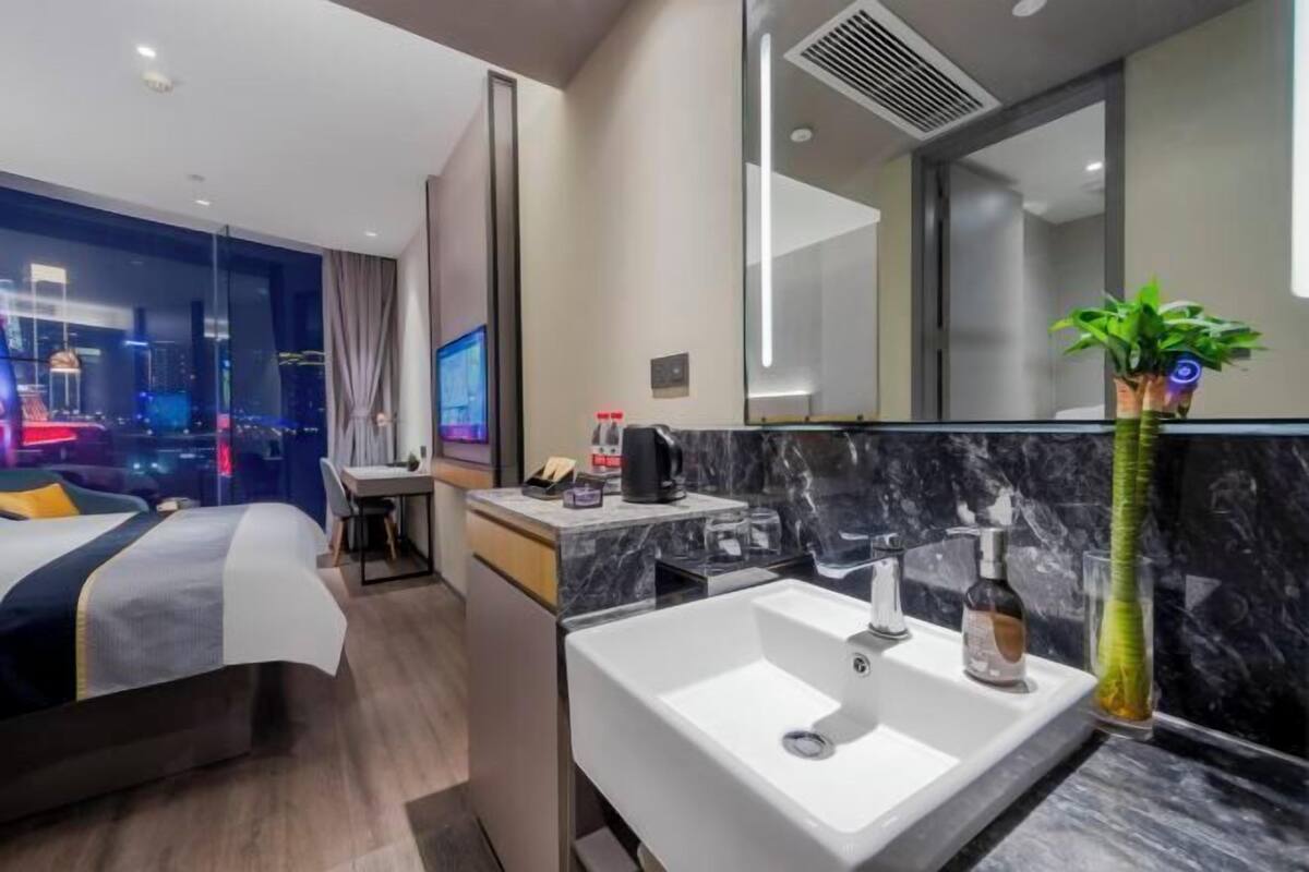 A modern hotel room with a bed, a desk, a bathroom with a sink, and a large mirror.