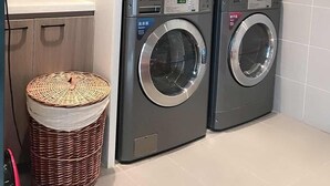 Un lavadero con dos lavadoras de carga frontal, una cesta de mimbre y un estante con libros y plantas.