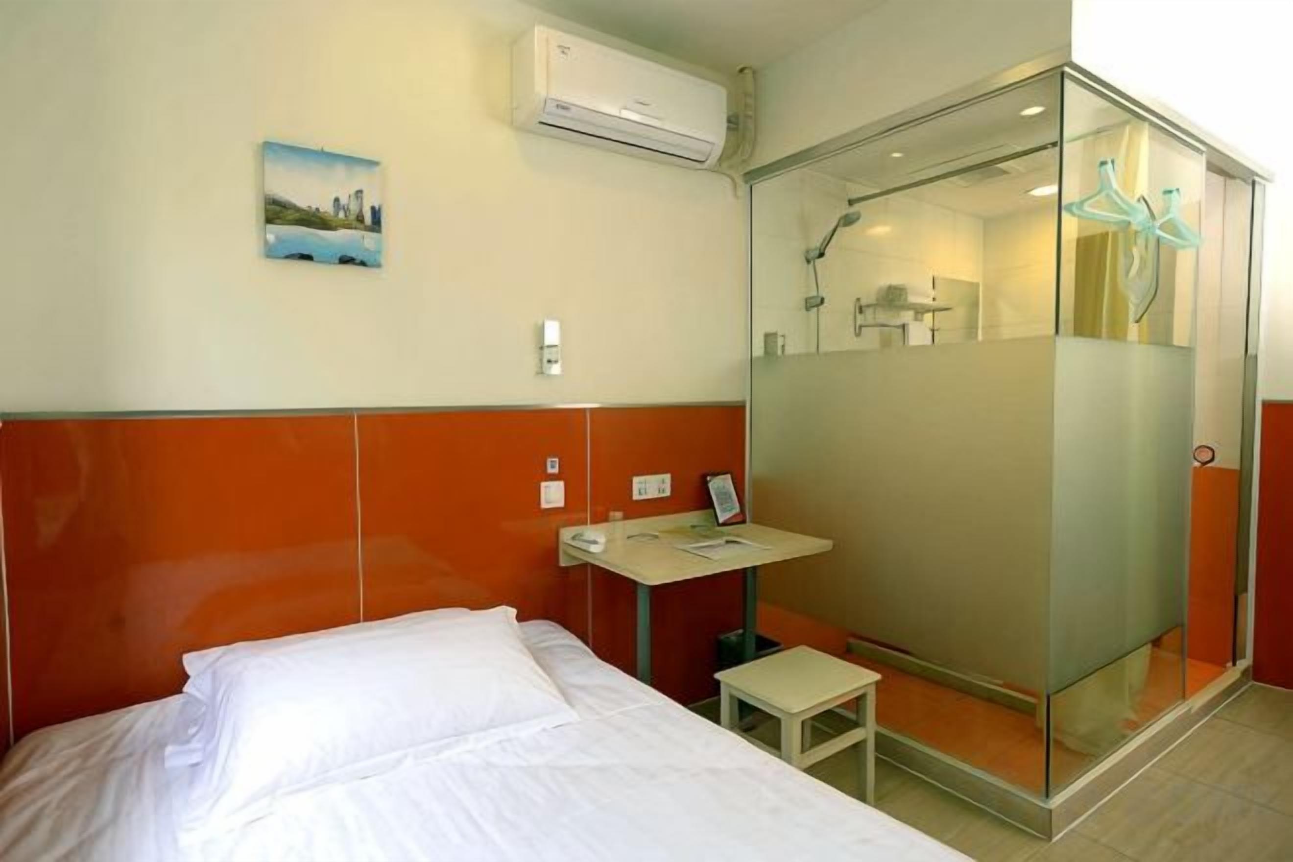 Habitación de hotel con cama, mesita de noche, espejo, equipo de aire acondicionado y cabina de ducha con mampara de vidrio.