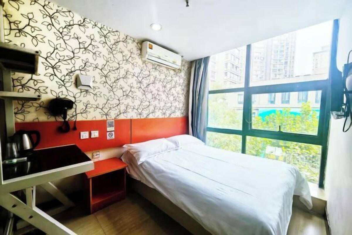 A compact hotel room with a single bed, a kitchenette, and a large window offering a view of buildings and greenery.