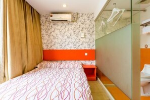 Habitación de hotel pequeña y funcional con cama, una mesita, unidad de aire acondicionado y cabina de ducha de vidrio.