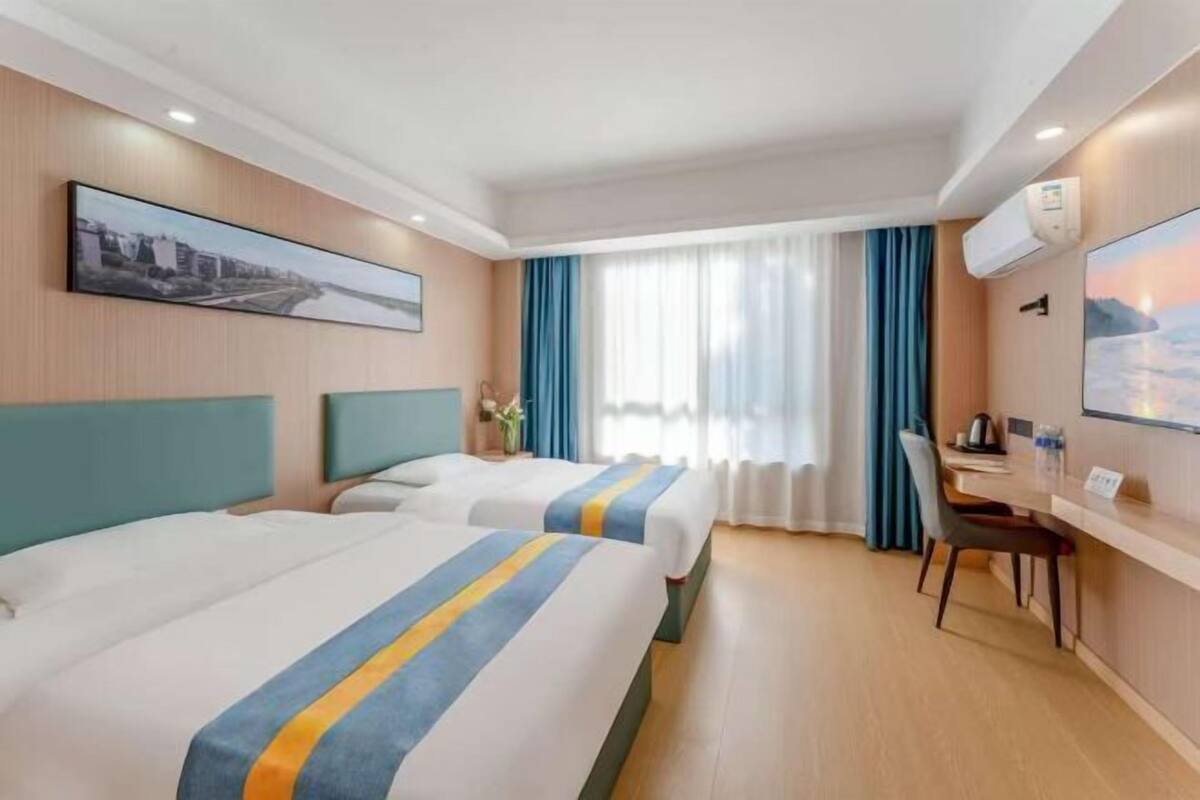 A hotel room with two beds, a desk, a chair, a window with curtains, and a wall-mounted air conditioner.
