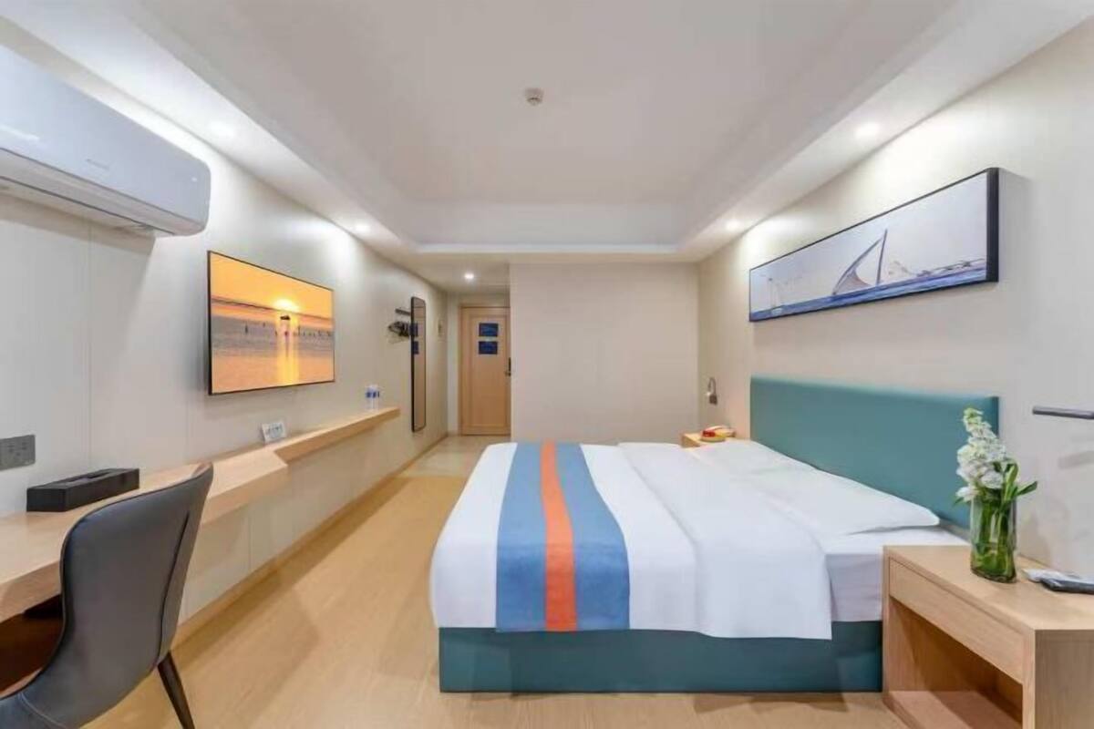 A hotel room with a bed, a desk, a chair, a nightstand, a vase with flowers, and a painting on the wall.