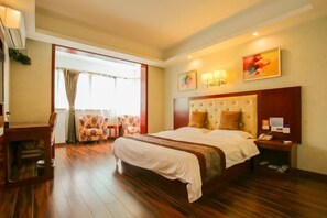 Une chambre d’hôtel avec un grand lit, un bureau et une chaise. Il y a une fenêtre avec des rideaux, un tableau au mur et une table de chevet avec une lampe.