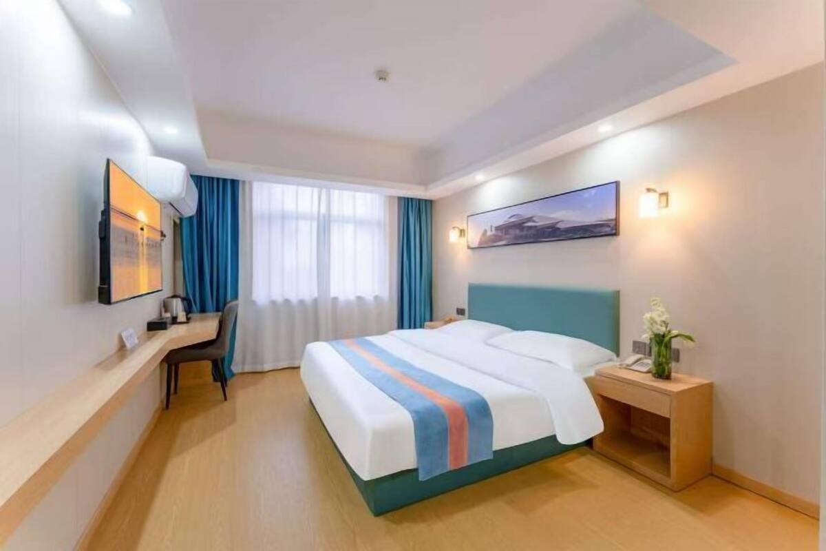 A hotel room with a bed, a desk, a chair, a nightstand, a picture on the wall, and a window with curtains.