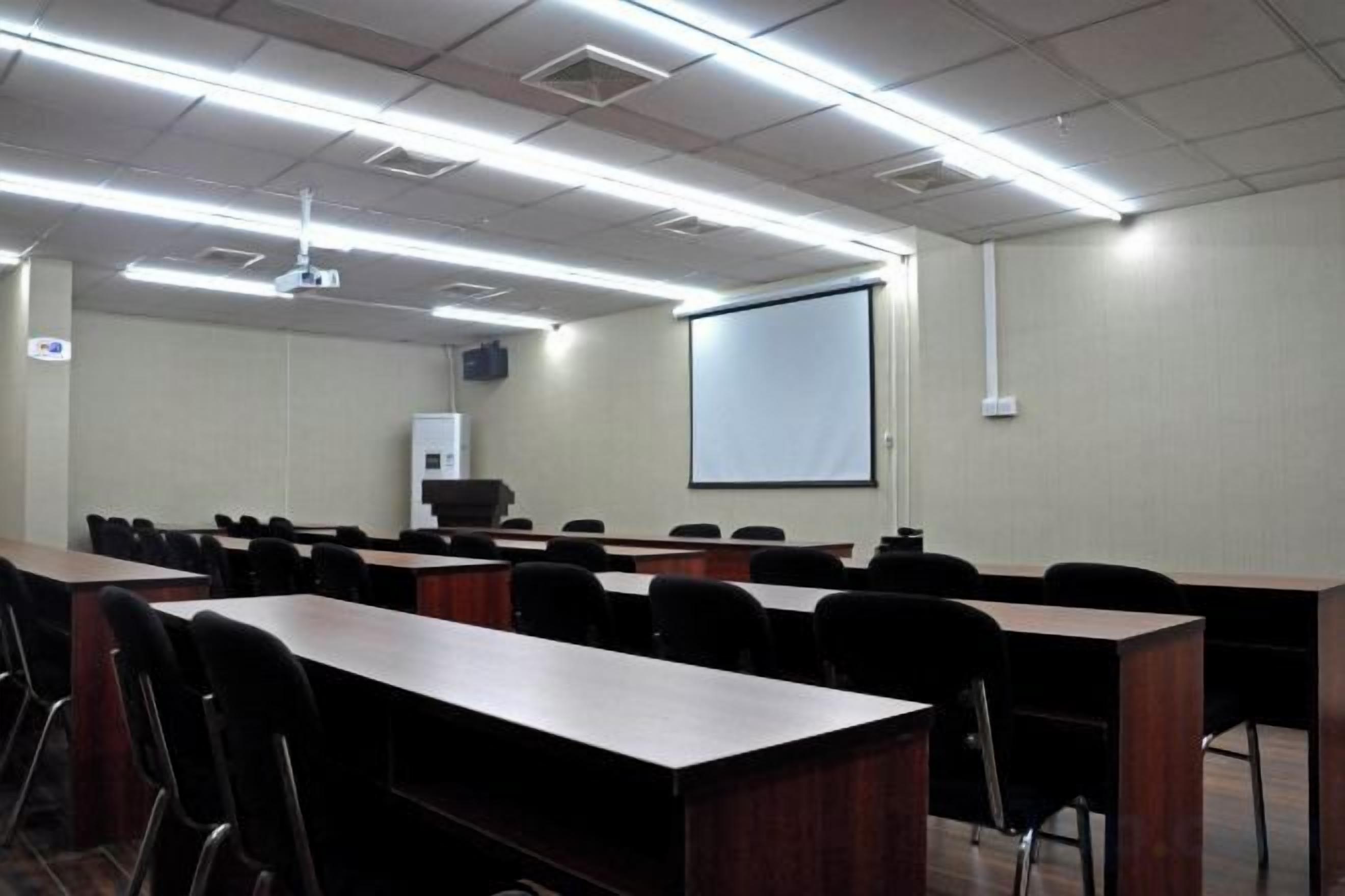 Un salón de conferencias con filas de mesas y sillas, una pantalla de proyector y una pizarra.