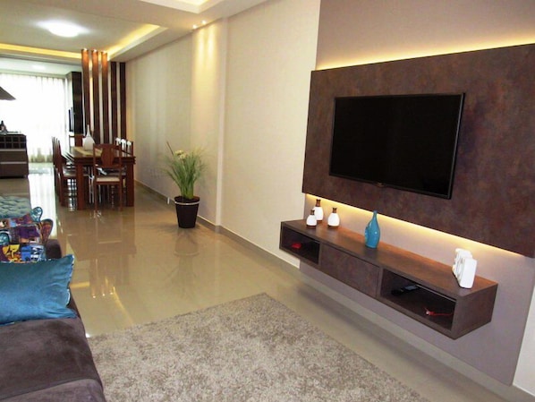 A modern living room with a flat-screen TV mounted on the wall, a dining area with chairs and a table, and a potted plant.