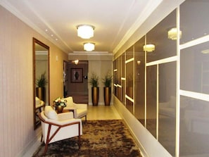 A hallway with a carpeted floor, two white armchairs, a small table with a vase of flowers, and tall planters.