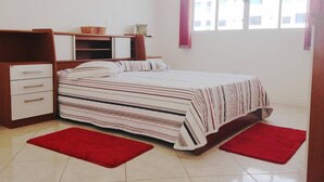 A bedroom with a bed, a desk, a chair, a red rug, and a window with a view of buildings. - Apartamento a 200m do Mar BCHost 17 (Itapema)