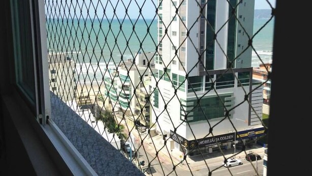 A view through a chain-link fence of a coastal cityscape with modern buildings, including a prominent white and green structure labeled "MOBILIÁNIA GOLDEN". - Apartamento a 200m do Mar BCHost 17 (Itapema)