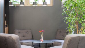 Une pièce avec deux fauteuils gris à revêtement en tissu disposés autour d’une petite table ronde sur laquelle se trouve un vase de fleurs. Des plantes en pot sont posées sur une étagère au-dessus de la table, et une grande plante se trouve à droite.