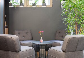Une pièce avec deux fauteuils gris à revêtement en tissu disposés autour d’une petite table ronde sur laquelle se trouve un vase de fleurs. Des plantes en pot sont posées sur une étagère au-dessus de la table, et une grande plante se trouve à droite.