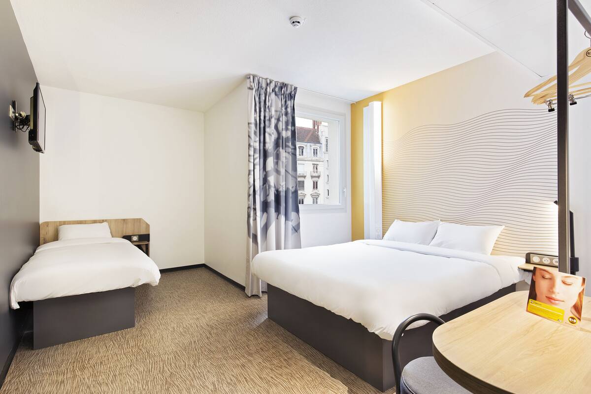 Een hotelkamer met twee bedden, een tafeltje, een stoel, een raam met gordijnen en een aan de muur bevestigde televisie.