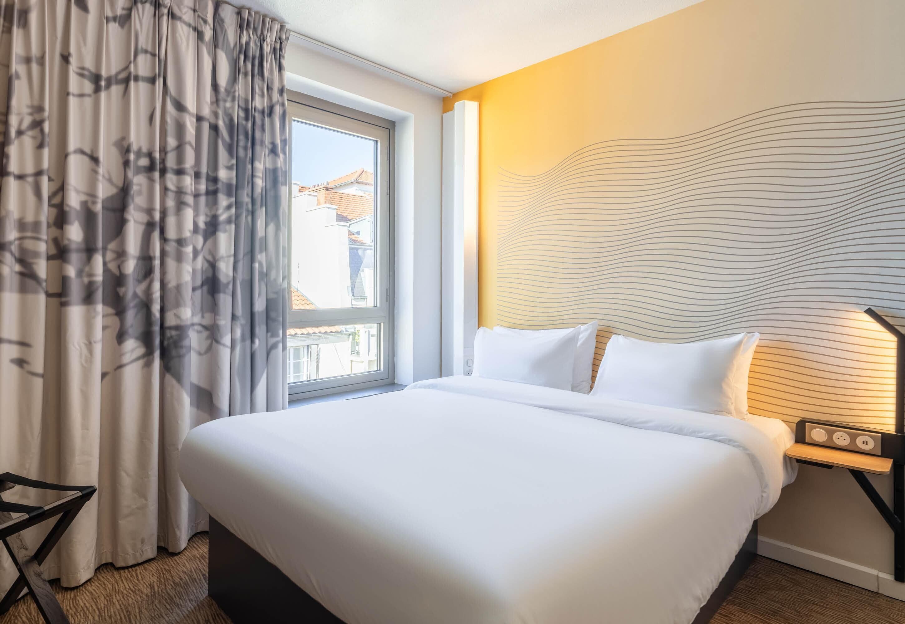 Habitación de hotel con una cama grande, una ventana con cortinas, una mesita de noche con una lámpara y una pared con diseño ondulado.