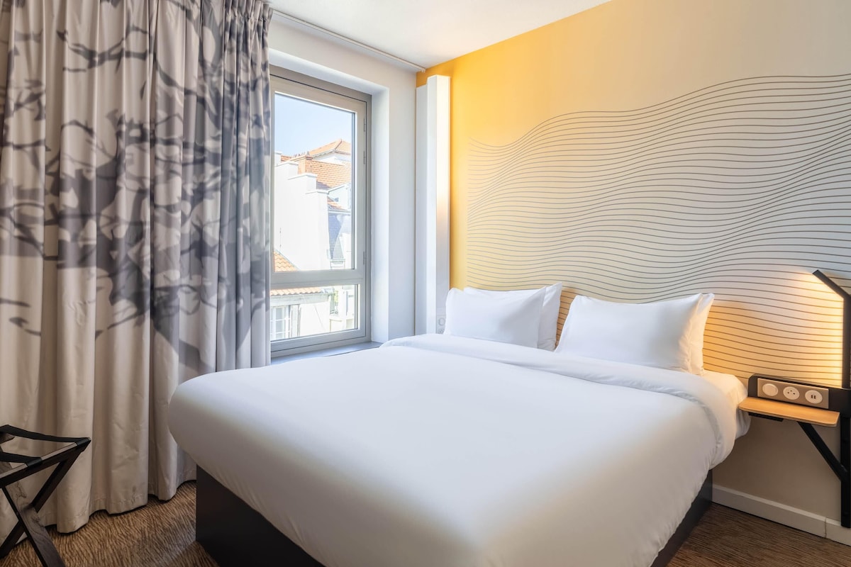 Habitación de hotel con una cama grande, una ventana con cortinas, una mesita de noche con una lámpara y una pared con diseño ondulado.
