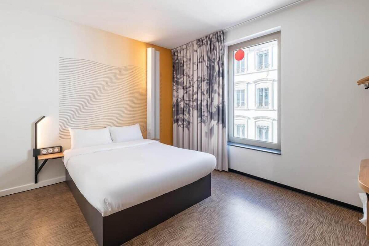Hotelkamer met een bed, nachtkastje, lamp, raam met gordijnen en uitzicht op gebouwen.
