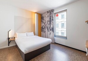 A hotel room with a bed, a nightstand, a lamp, a window with curtains, and a view of buildings. - B&B HOTEL LYON Centre Perrache (Lyon)
