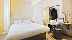 Une chambre d’hôtel moderne équipée d’un grand lit, d’un bureau avec un ordinateur, d’une armoire et d’un miroir.