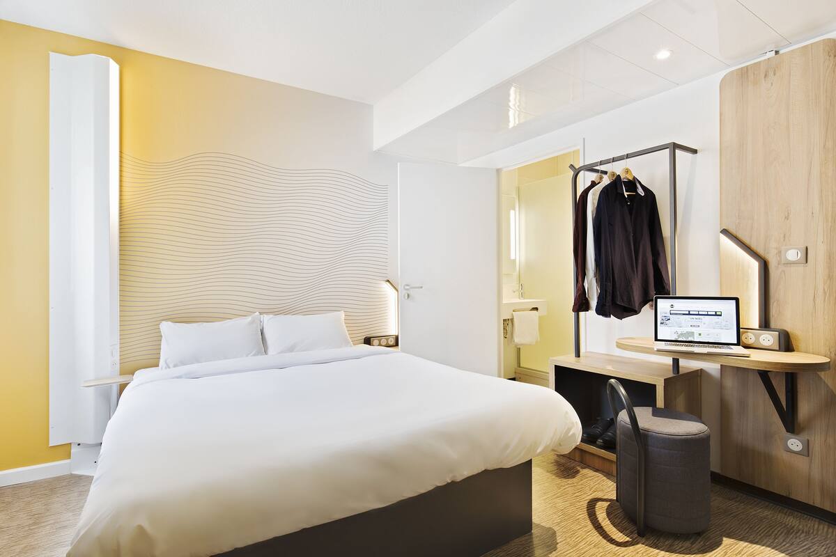 Een moderne hotelkamer met een bed, een bureau met een computer, een stoel, een kapstok en een prullenbak.