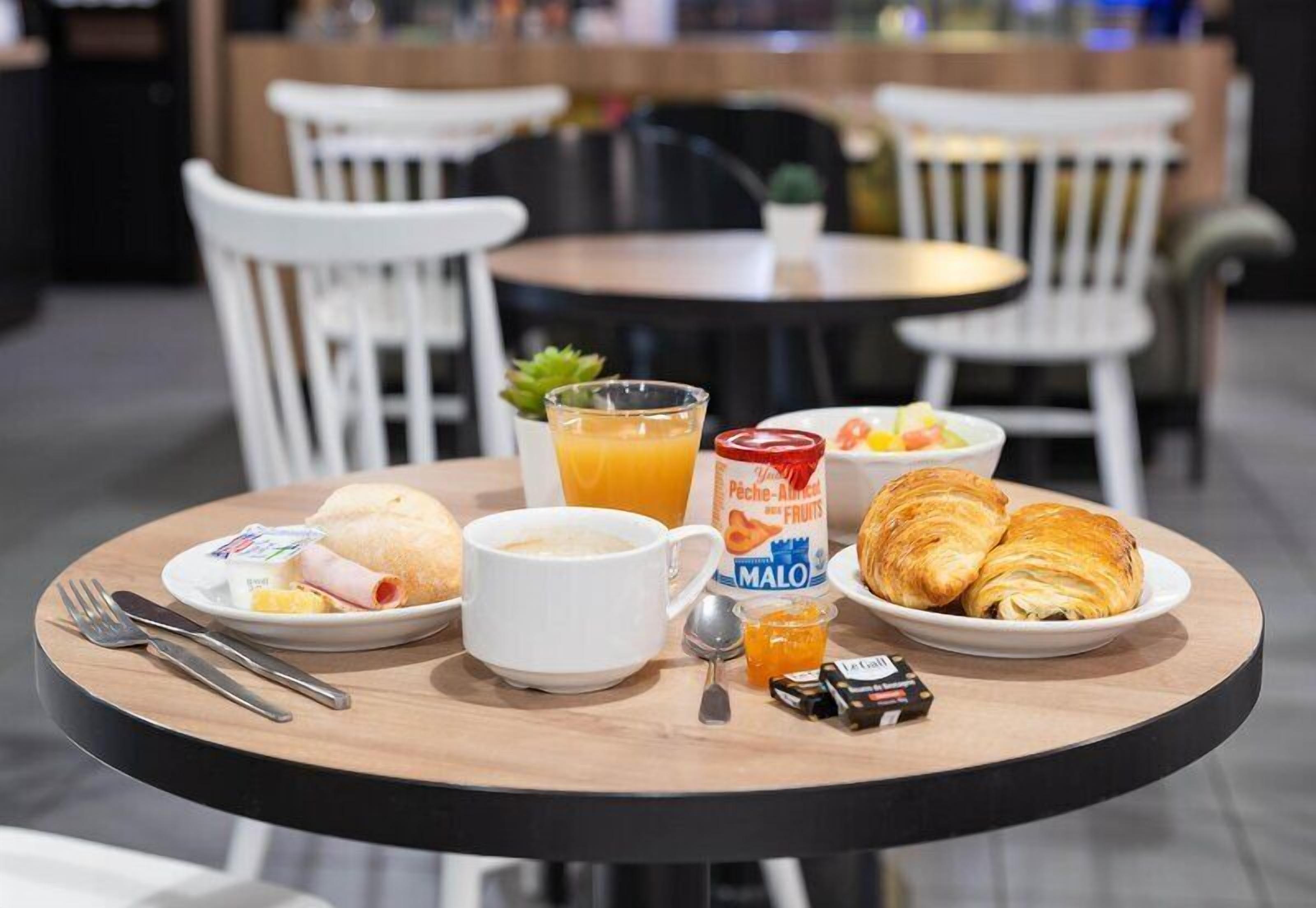 Una mesa de desayuno con croissants, un frutero, un vaso de zumo de naranja, una taza de café y un plato con un pastel.