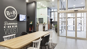 Un hall d’hôtel moderne avec un comptoir d’accueil en bois, un téléviseur affichant des images de la chambre et une entrée avec une porte en verre.