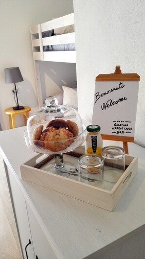 A breakfast tray with pastries, a glass of milk, and a small bottle of jam on a wooden table. A welcome sign with the text "Benvenuti Welcome" and "Quartino Napoli" is visible. - Quartino Napoletano (Naples)