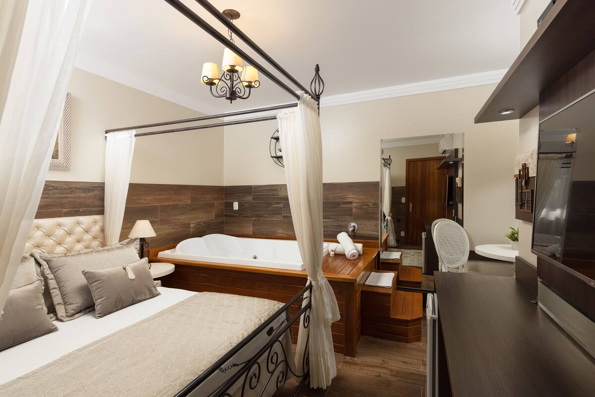 A bedroom with a bed, a sitting area with a chair, a vanity with a mirror, and a bathtub.