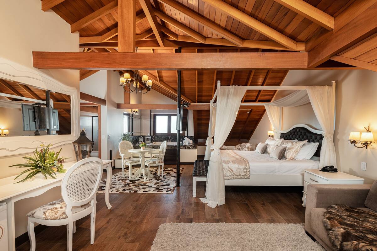 A bedroom with a canopy bed, wooden beams, a desk with a chair, and a dining area.