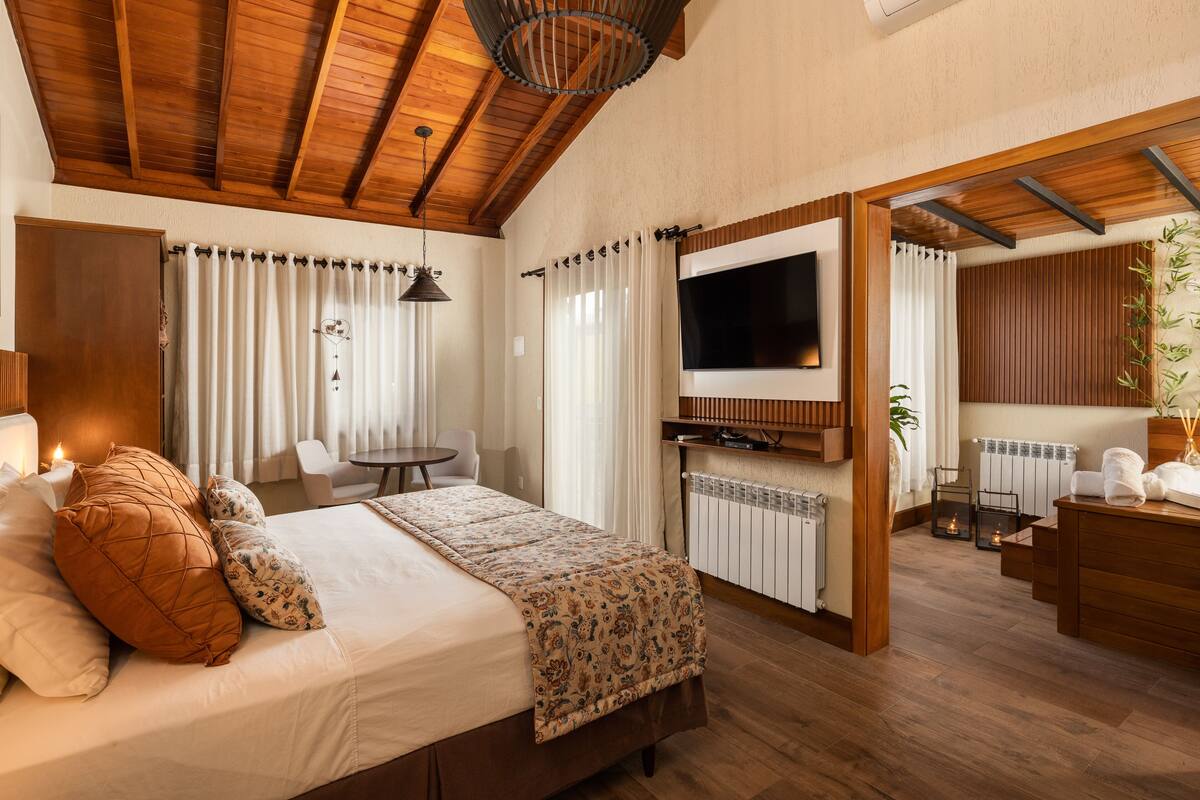 A hotel room with a wooden ceiling, a bed with a patterned bedspread, a TV mounted on a wooden panel, a small table with a chair, and a radiator.