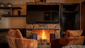 Una sala de estar acogedora con una chimenea de piedra, dos sillones de terciopelo, un televisor de pantalla plana sobre la chimenea y una estantería con libros y objetos decorativos.