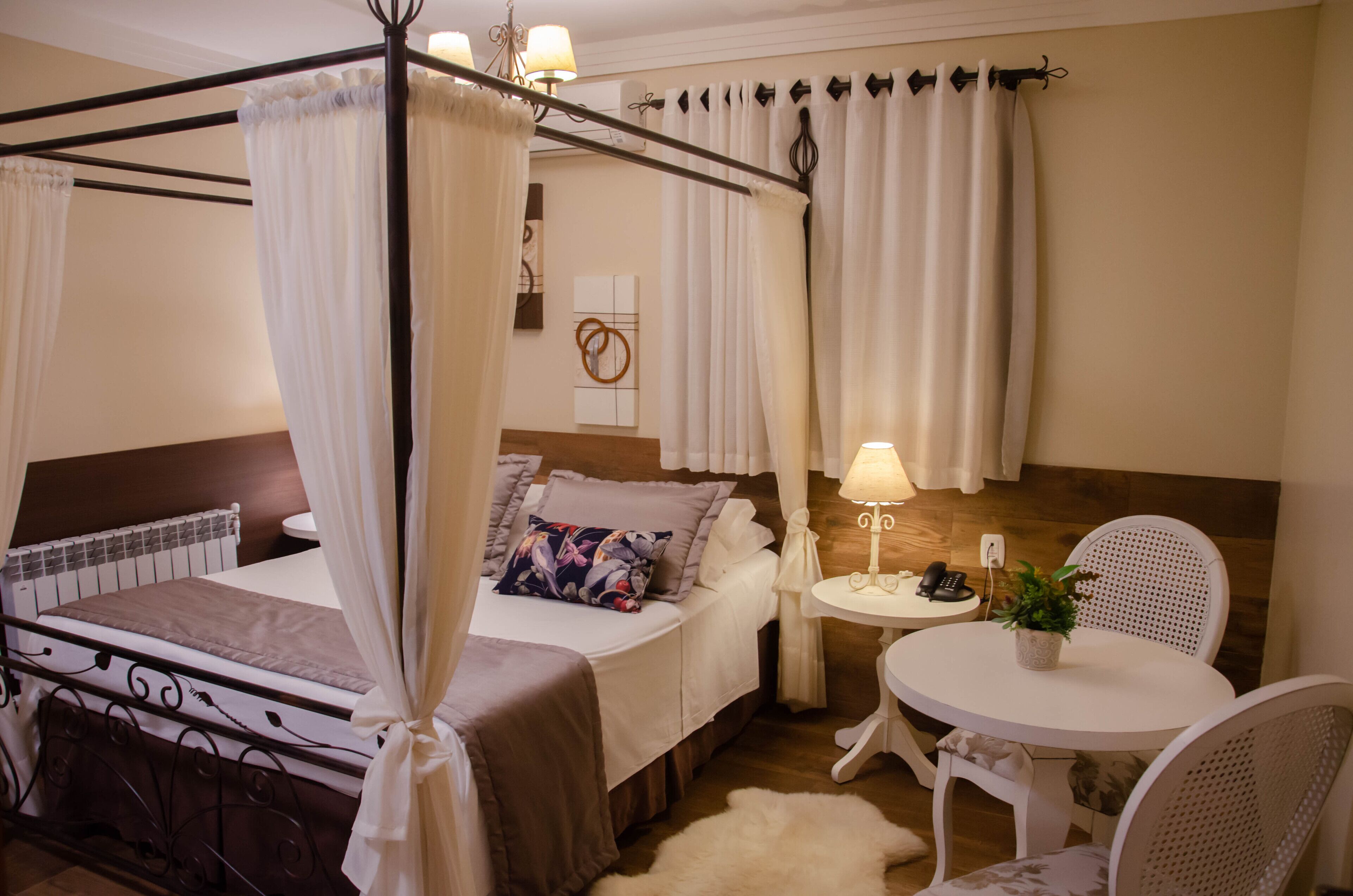 Un dormitorio con cama con dosel, una mesita de noche con lámpara, una pequeña mesa redonda con sillas y una planta en maceta.