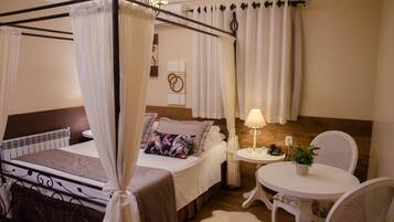 Un dormitorio con cama con dosel, una mesita de noche con lámpara, una pequeña mesa redonda con sillas y una planta en maceta.