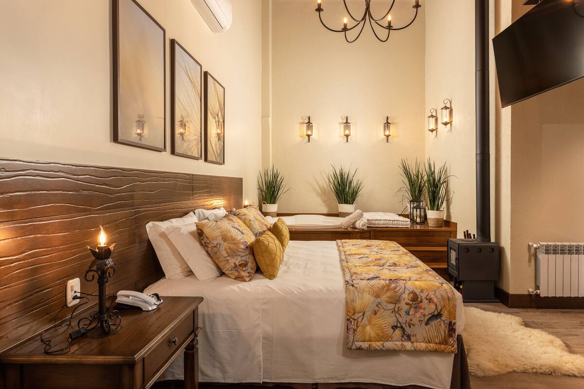 A bedroom with a wooden headboard, a bedside table with a lamp, a bed with floral bedding, a wall-mounted TV, and a fireplace.