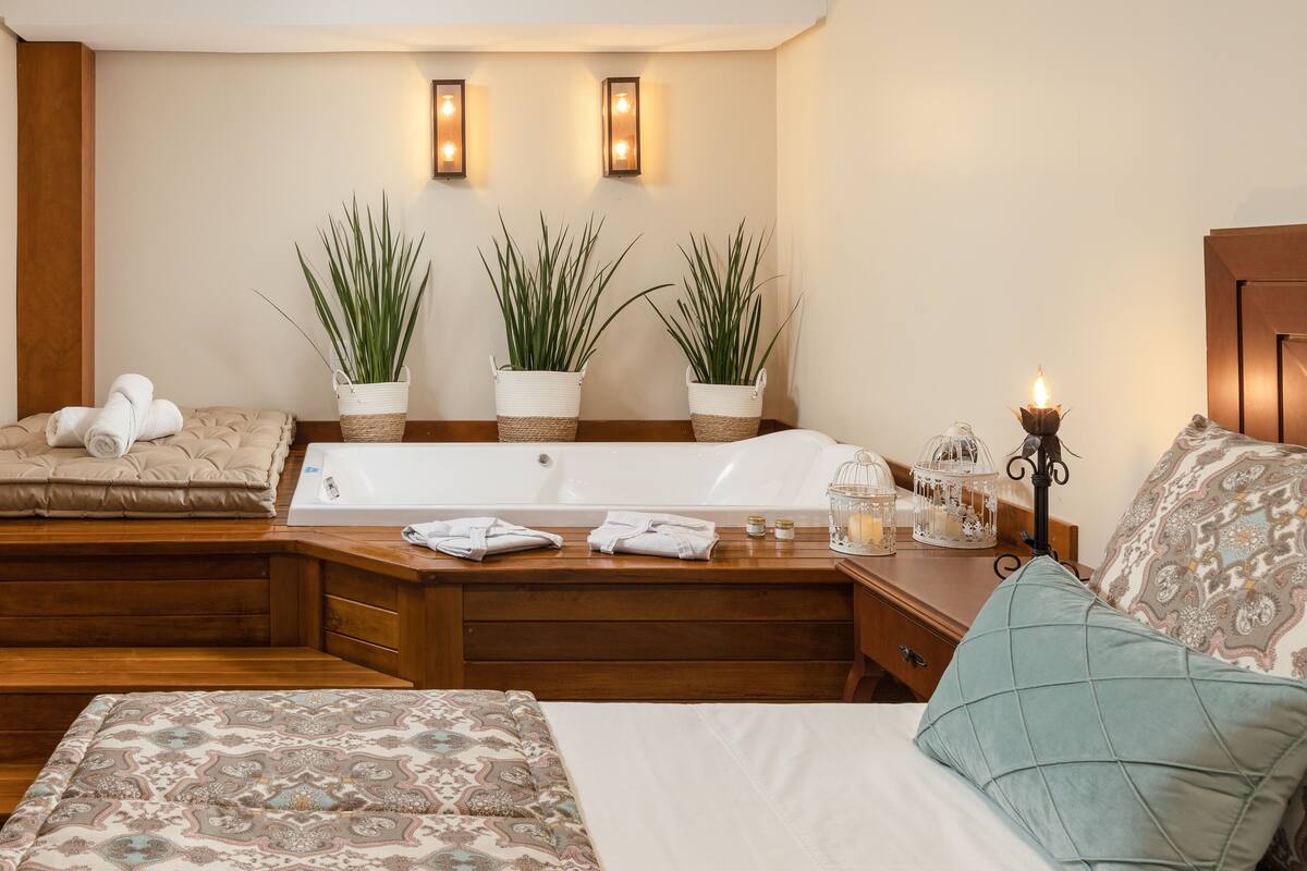 A modern bedroom with a large bed, a wooden nightstand, a built-in bathtub, and two potted plants.