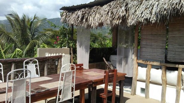 An outdoor dining area with a wooden table and chairs, a thatched roof, and a view of greenery and mountains. - Le Creole Hideaway (Jacmel)