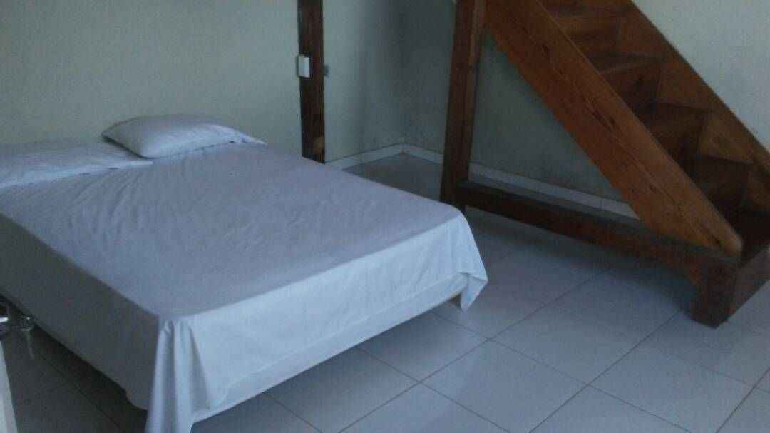 Uma cama de solteiro com lençol branco num quarto com chão de azulejos e escada de madeira.