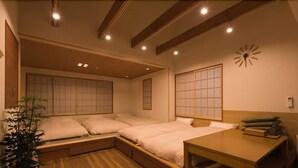 A traditional Japanese-style bedroom with a low ceiling, wooden beams, sliding shoji screens, a bed with a wooden headboard, a bedside table, and a potted plant. - Guestvilla Hakone Miyanoshita (Hakone)