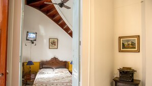 A bedroom with a wooden ceiling fan, a bed with floral bedding, a nightstand with a lamp, and a small desk with a typewriter. - Rio Antigo (Rio de Janeiro)