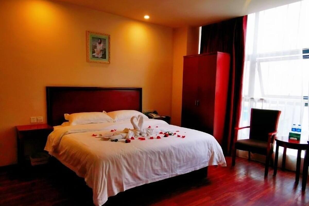 A hotel room with a large bed, a wooden floor, a nightstand, a chair, and a wardrobe.