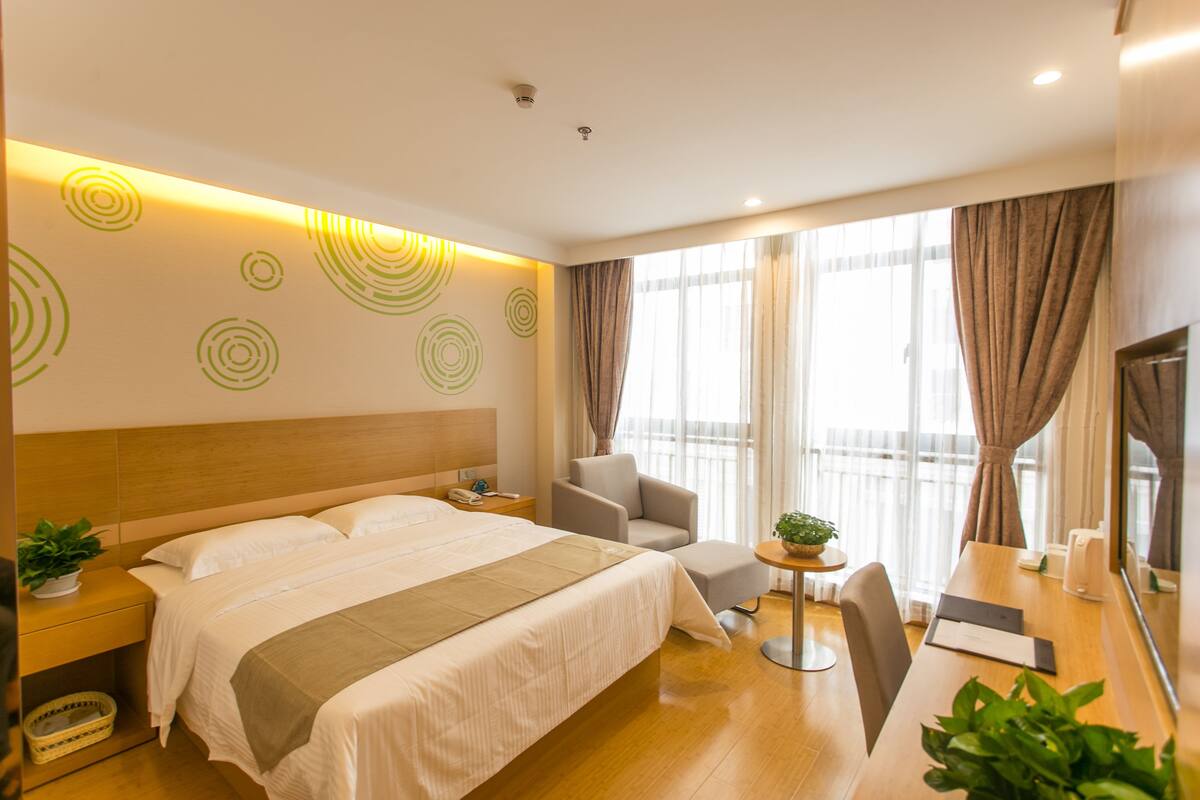 Ein Hotelzimmer mit einem großen Bett, einem Schreibtisch, einem Stuhl, einem kleinen Tisch mit Pflanze und einem Fernseher.