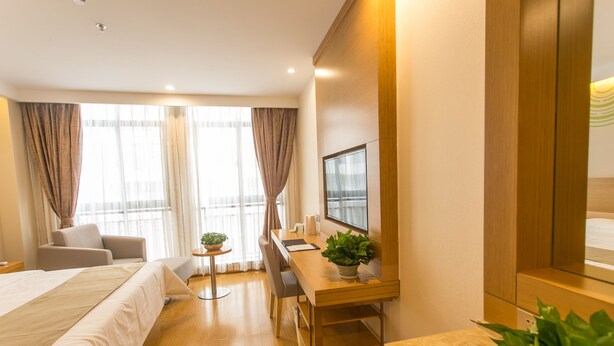 A hotel room with a bed, a desk, a chair, a small table with a plant, and a TV mounted on the wall.