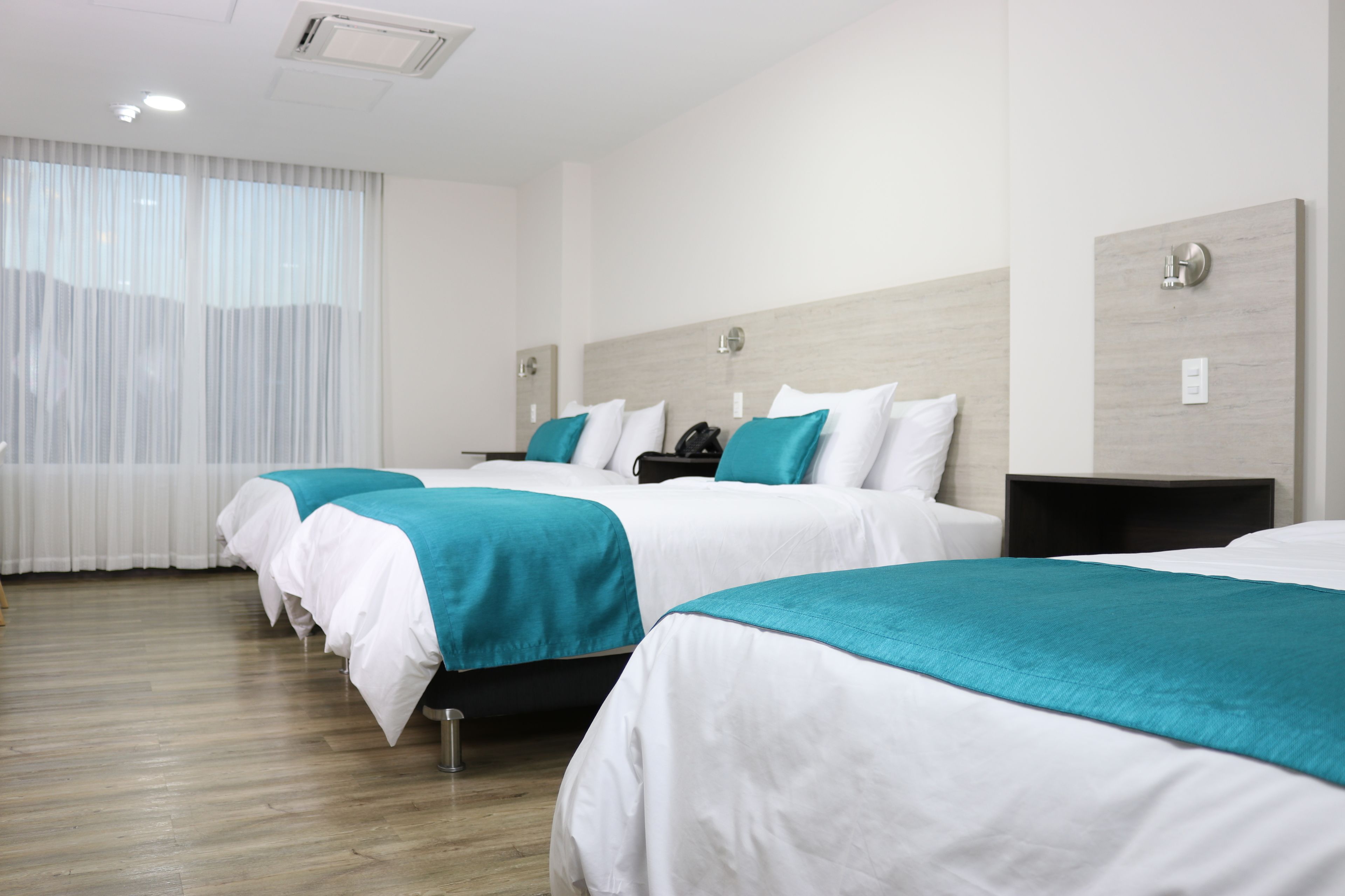 Habitación de hotel con tres camas, cada una con ropa de cama blanca y almohadones turquesa. El suelo es de madera, hay una ventana con cortinas traslúcidas y una lámpara de pared.