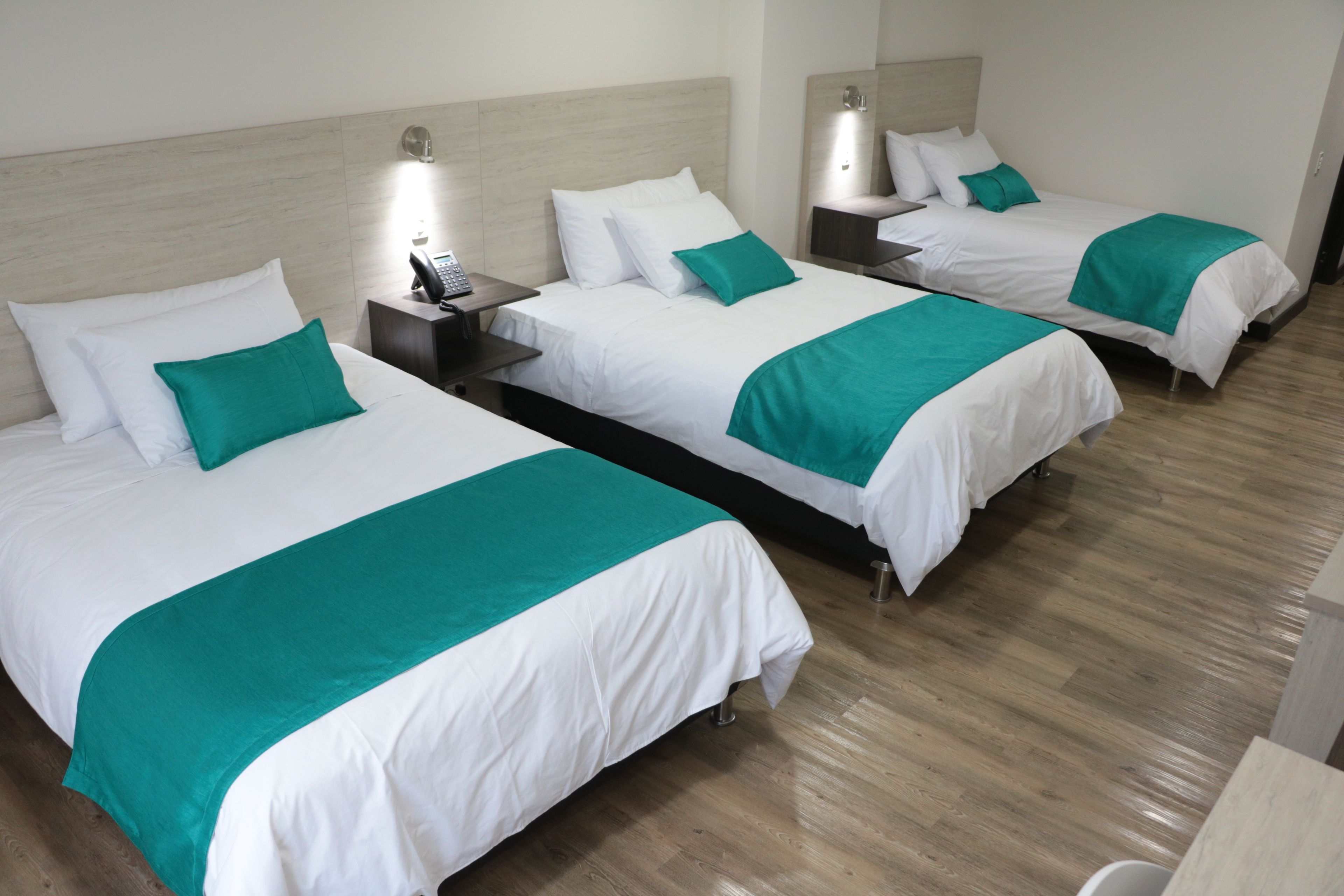 Une chambre d’hôtel avec trois lits, chacun doté de draps blancs et agrémenté de touches de couleur turquoise. On y trouve des tables de chevet avec des lampes et un téléphone.