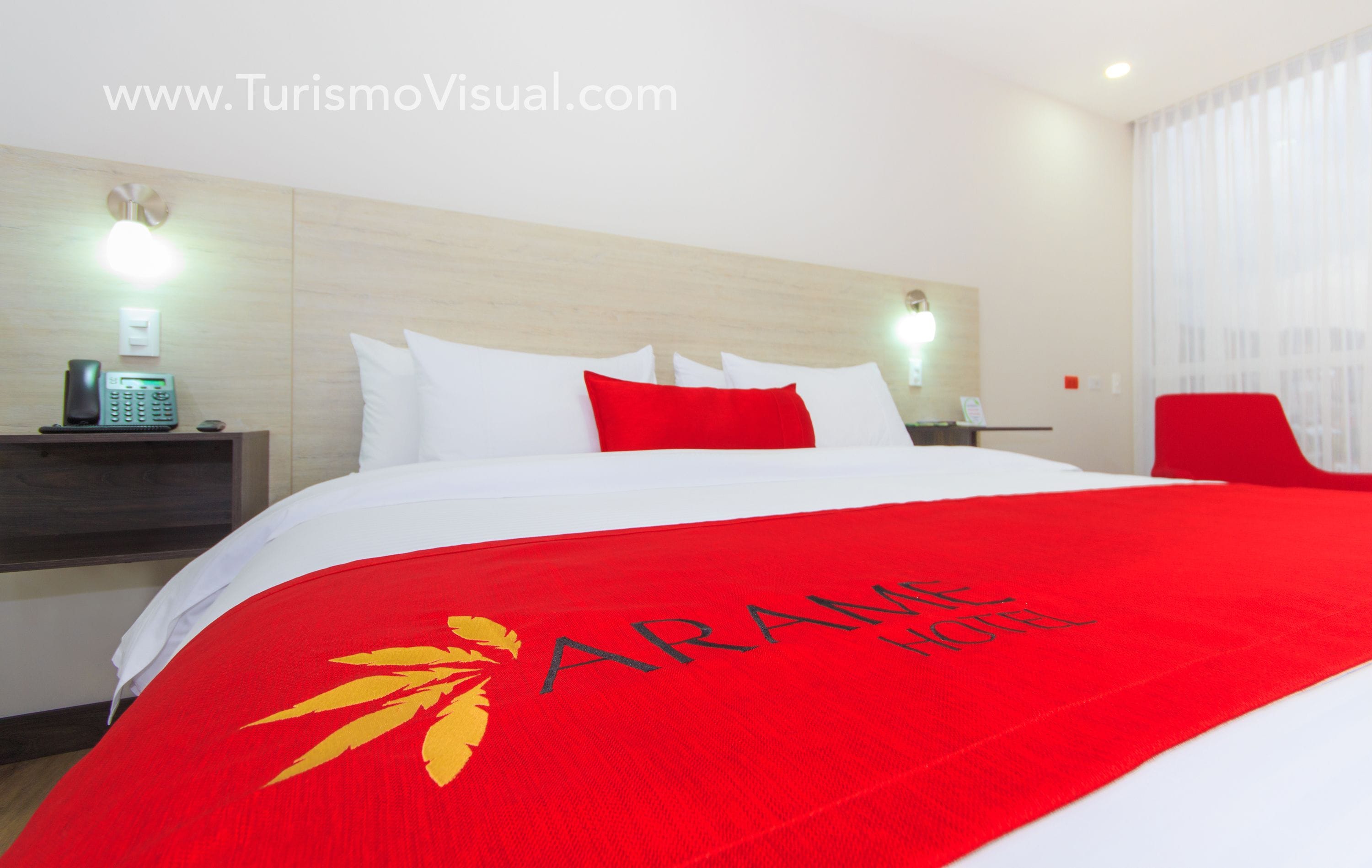A hotel room with a large bed, red and white bedding, a red chair, and a nightstand with a phone.