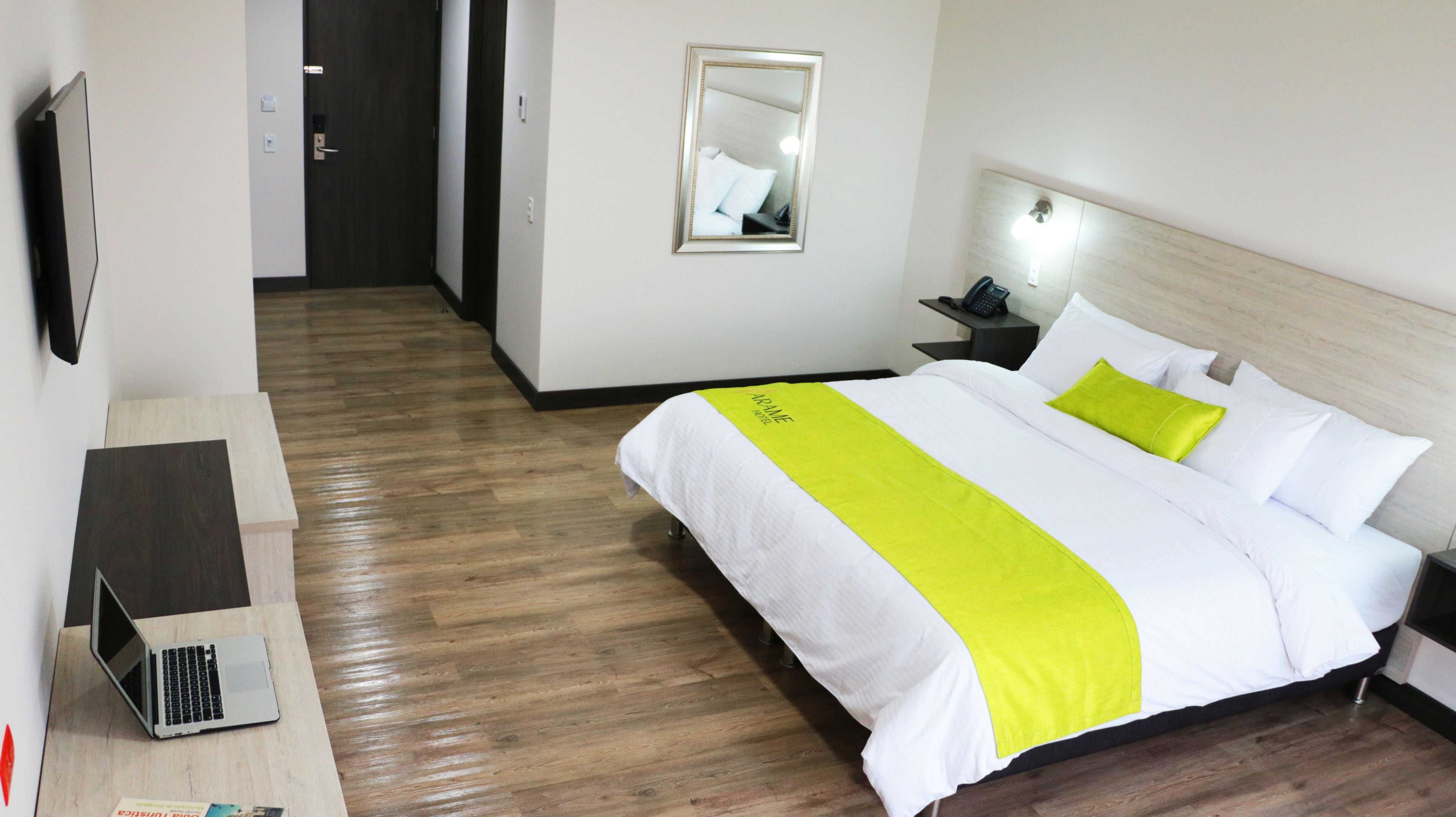 A hotel room with a bed, a desk with a laptop, a bedside table with a phone, and a mirror on the wall.