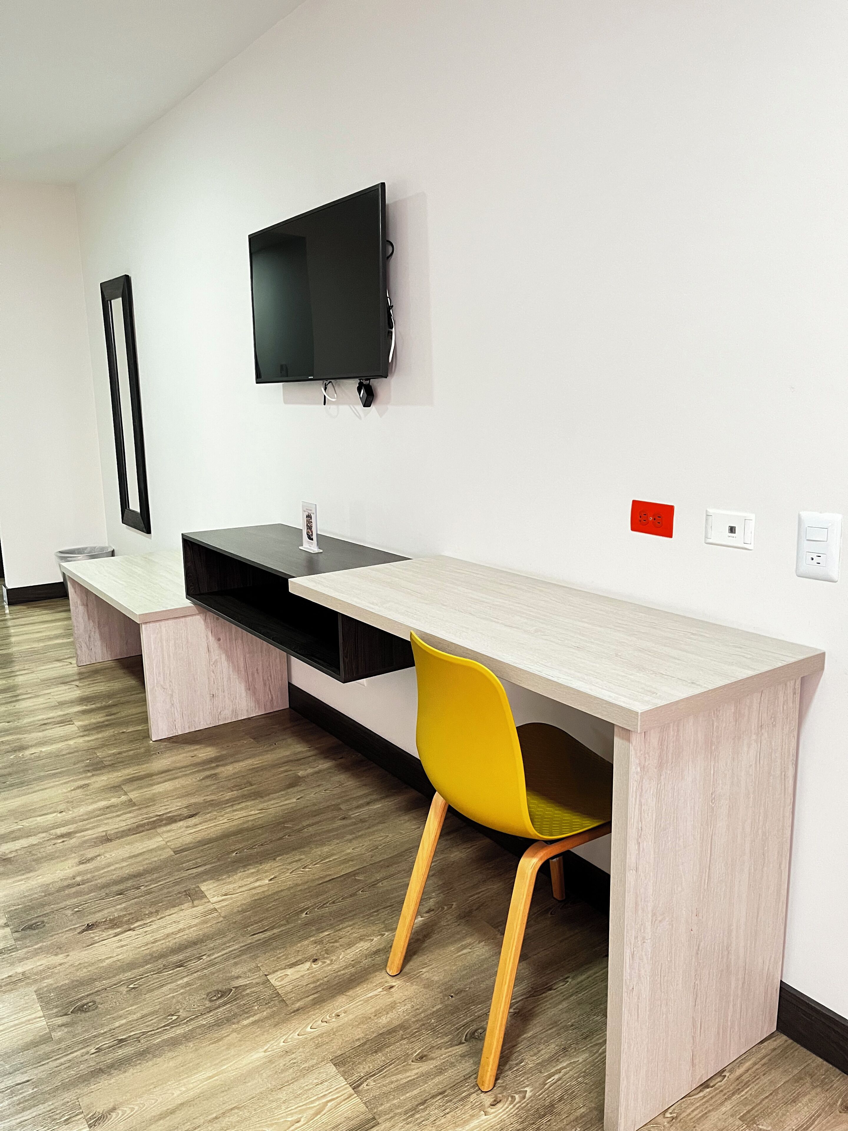 Une pièce moderne et minimaliste avec un plancher en bois, un grand bureau agrémenté d’une chaise jaune, un téléviseur noir fixé au mur et une petite prise électrique rouge et blanche.
